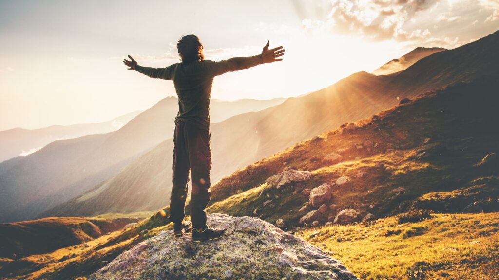 Man with arms stretched out in surrender toward heaven standing on a mountain and looking at the sun.